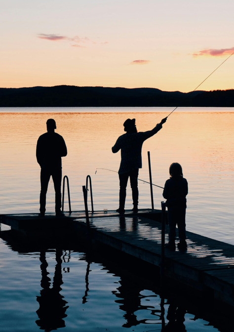 Kolme kalastajan hahmoa laiturilla, tyyni järvi, oransi taivaanranta.