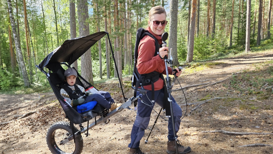 Kuvassa nainen kulkee nousevaa metsäpolkua vetäen Cascade-maastokärryssä alle kouluikäistä lasta.