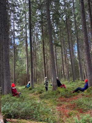 Ihmisiä metsässä istuskelemassa ja nojailemassa puihin.