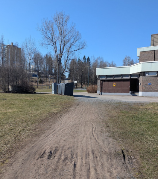 Nurmen rajaama hiekkaväylä. Väylällä pyörän tekemiä uria ja kuoppia. Edessäpäin puita ja rakennuksia.