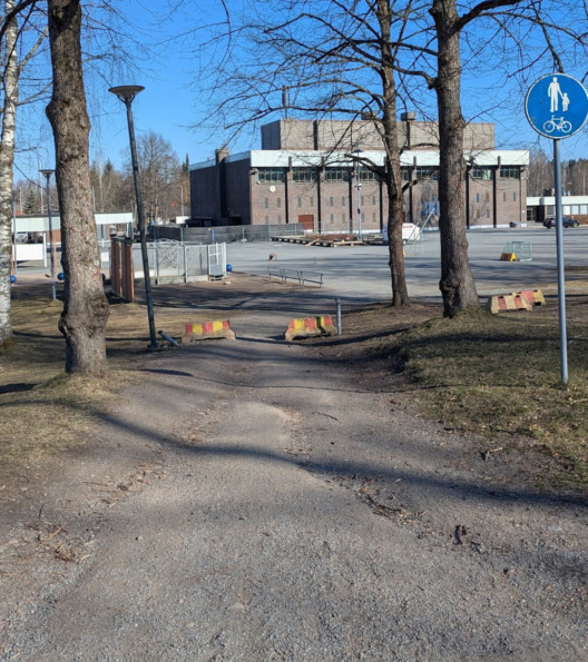 Hiekkaväylä laskeutuu mäkeä alas. Kulkuväylällä sadeveden syövyttämiä kohtia. Edessä puita ja rakennuksia.