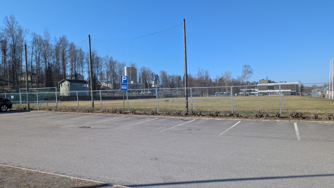 Asfalttipintainen parkkialue, jossa paikat ovat vieretysten. Parkkialueen edessä verkkoaita ja jalkapallokenttä.  