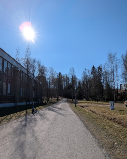 Nurmen rajaamaa asfalttiväylää. Vasemmalla puolella punatiilistä tehty kaksikerroksinen rakennus. Edessä puita ja asfaltissa näkyy halkeamia. 